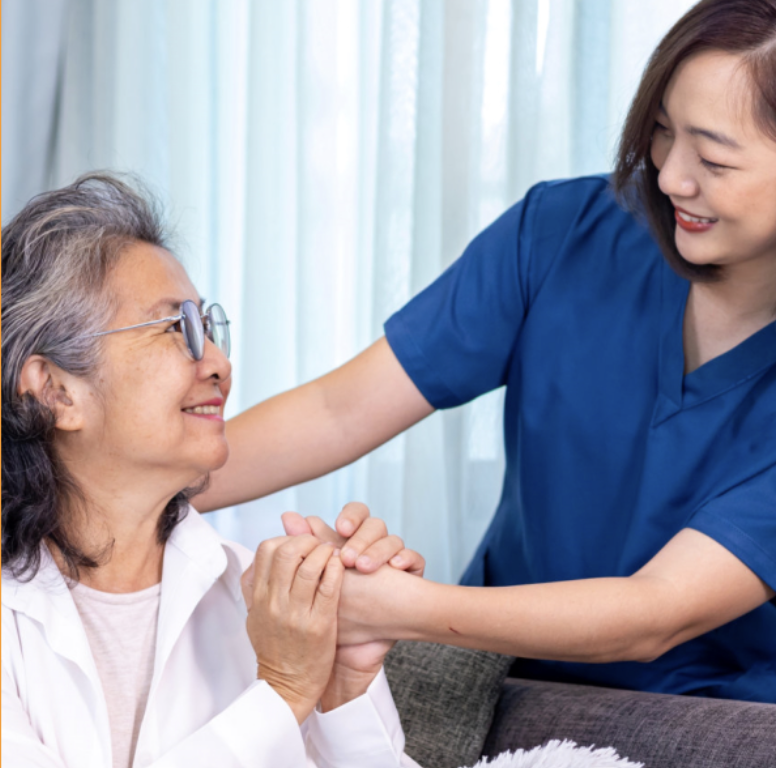 Caregiver in blue uniform gently holding hands with an older woman in a white shirt, both seated in a comfortable home setting with soft natural light and a calm, supportive atmosphere in Huntersville, NC
